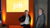 Zwei Personen stehen in einem Raum und unterhalten sich. Im Hintergrund ist ein orangefarbenes Banner mit dem Schriftzug "srh" zu sehen.