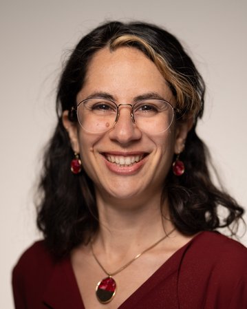 Eine lächelnde Person mit dunklen, lockigen Haaren trägt eine rote Bluse und Schmuck. Sie hat eine Brille und steht vor einem neutralen Hintergrund.