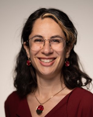 Eine lächelnde Person mit dunklen, lockigen Haaren trägt eine rote Bluse und Schmuck. Sie hat eine Brille und steht vor einem neutralen Hintergrund.