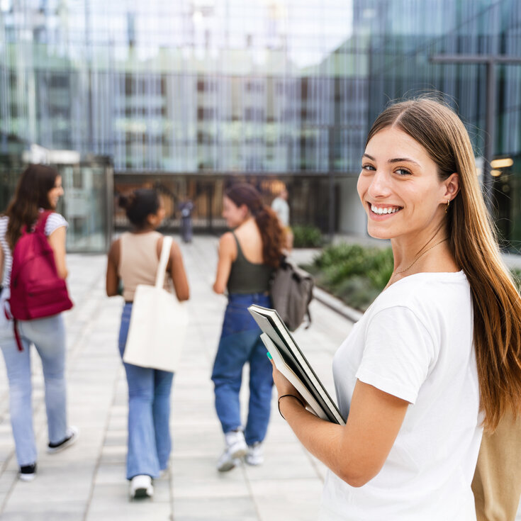Eine junge Frau mit langen Haaren hält Bücher in der Hand und lächelt, während sie einer Gruppe von drei anderen Frauen in einem modernen Umfeld folgt.