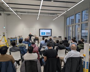 A group of people is sitting in a modern room, listening to a presentation. A speaker stands in front of a screen displaying a slideshow. Some technical equipment is visible in the background.