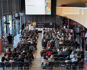 A large crowd is seated in a modern event space while a speaker is giving a presentation on stage.