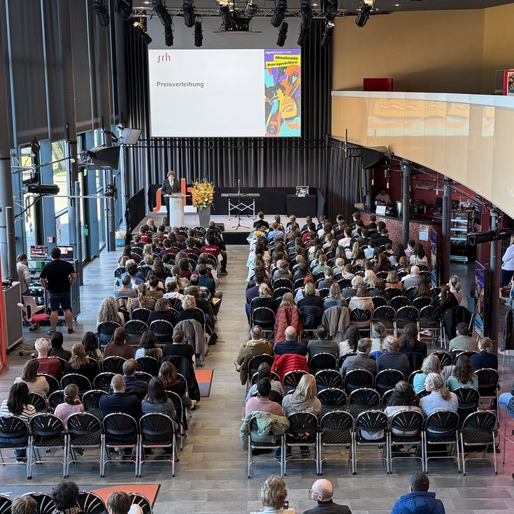 A large crowd is seated in a modern event space while a speaker is giving a presentation on stage.