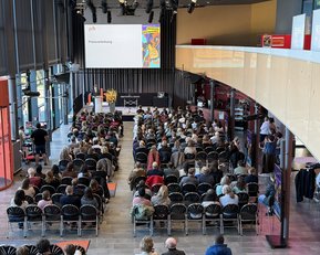 A large crowd is seated in a modern event space while a speaker is giving a presentation on stage.