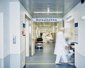 Flur in einem Krankenhaus mit dem Schild "Notaufnahme". Ein Arzt und Patienten sind sichtbar, während ein Rollstuhl vorbeigeschoben wird.