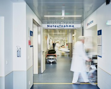 Flur in einem Krankenhaus mit dem Schild "Notaufnahme". Ein Arzt und Patienten sind sichtbar, während ein Rollstuhl vorbeigeschoben wird.