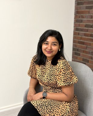 Eine Person mit schwarzem Haar sitzt in einem Sessel und trägt ein gepunktetes Kleid. Sie lächelt und schaut direkt in die Kamera.