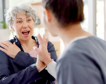 Eine ältere Frau mit grauen Haaren zeigt Freude und Überraschung im Gespräch mit einer jüngeren Person in einem hellen Raum.