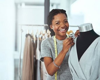 Eine lächelnde Frau mit Zopf frisiert ein Kleidungsstück an einer Schneiderpuppe, während sie ein Maßband um den Hals trägt.