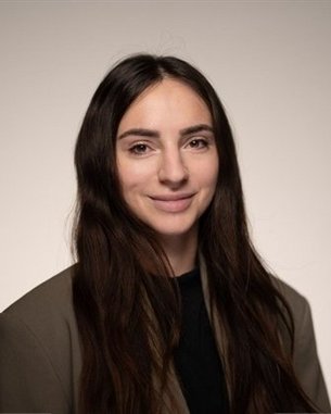 Eine Person mit langen, dunklen Haaren und einem neutralen Gesichtsausdruck trägt einen schwarzen Oberteil und einen grauen Blazer.