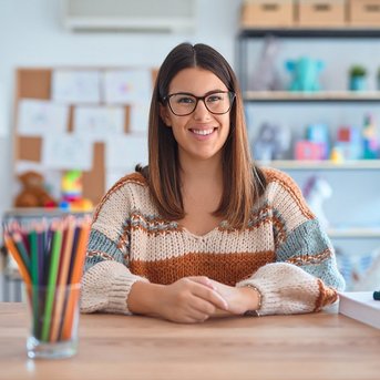 Eine junge Frau mit langen, braunen Haaren und Brille sitzt an einem Tisch. Sie trägt einen gestreiften Pullover und lächelt. Auf dem Tisch befinden sich Buntstifte, Spielzeug und andere Materialien.