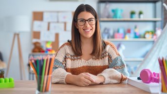Eine junge Frau mit langen, braunen Haaren und Brille sitzt an einem Tisch. Sie trägt einen gestreiften Pullover und lächelt. Auf dem Tisch befinden sich Buntstifte, Spielzeug und andere Materialien.