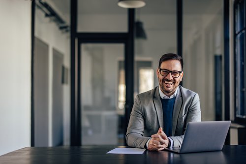 Ein Mann in einem grauen Anzug sitzt an einem Tisch mit einem Laptop und lächelt. Der Hintergrund zeigt ein modernes Büro.