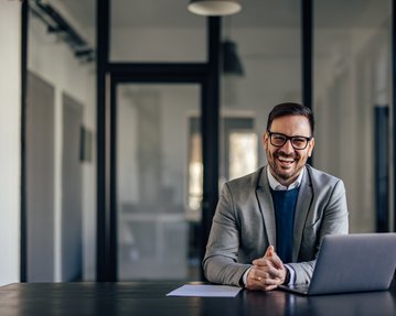 Ein Mann in einem grauen Anzug sitzt an einem Tisch mit einem Laptop und lächelt. Der Hintergrund zeigt ein modernes Büro.