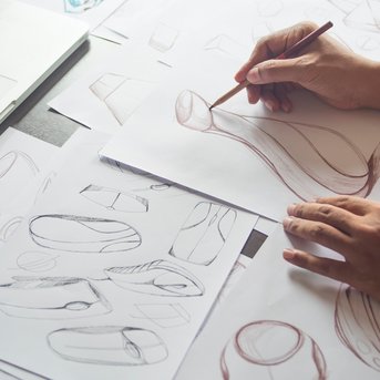 Hands sketch designs on paper, surrounded by additional drawings and a laptop on the table.