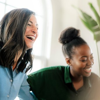 Drei Frauen lachen und interagieren in einem hellen Raum, umgeben von Pflanzen. Sie tragen lässige Kleidung und scheinen eine angenehme Zeit zu haben.