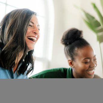 Three women are laughing and interacting in a bright room surrounded by plants. They are dressed casually and seem to be having a great time.