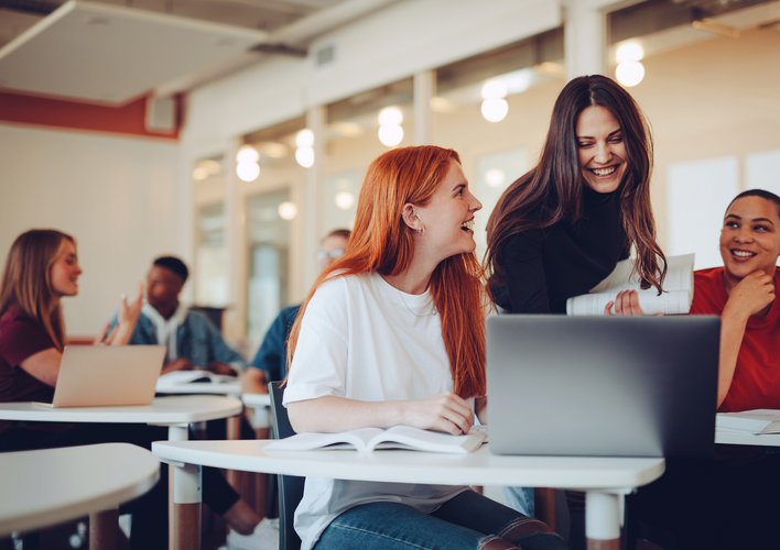 Eine Gruppe von Studierenden sitzt in einem modernen Klassenraum, einige arbeiten an Laptops, während andere miteinander sprechen und lachen.
