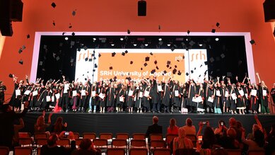 Graduates of SRH University Heidelberg celebrate their graduation by throwing their caps in the air while standing on stage.