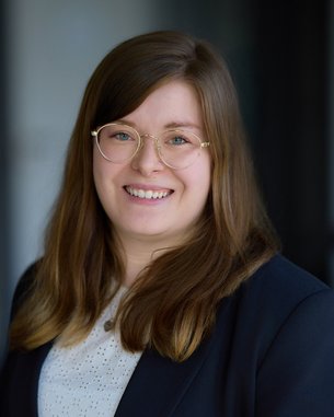 Eine lächelnde Frau mit langen, braunen Haaren und Brille trägt einen schwarzen Blazer und ein weißes Oberteil.