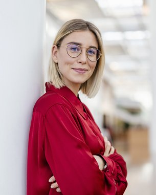 Eine junge Frau mit blonden, schulterlangen Haaren und Brille steht an einer Säule in einem modernen Innenraum. Sie trägt ein rotes Hemd und hat die Arme verschränkt. Der Hintergrund zeigt eine helle, offene Umgebung mit Pflanzen.