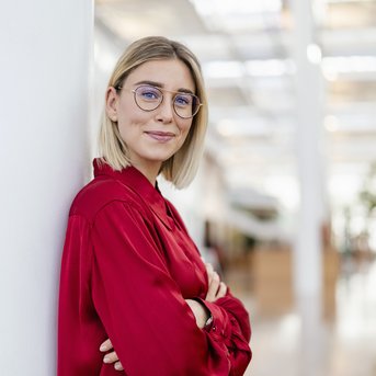 Eine junge Frau mit blonden, schulterlangen Haaren und Brille steht an einer Säule in einem modernen Innenraum. Sie trägt ein rotes Hemd und hat die Arme verschränkt. Der Hintergrund zeigt eine helle, offene Umgebung mit Pflanzen.