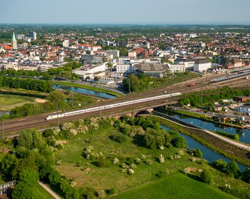 Luftaufnahme einer Stadt mit einem großen Bahngleis, das über einen Fluss führt. Im Hintergrund sind moderne Gebäude und grüne Landschaften zu sehen.