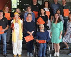 A group of 17 people is standing on a staircase, some holding orange documents. The participants are dressed in various styles and are smiling.