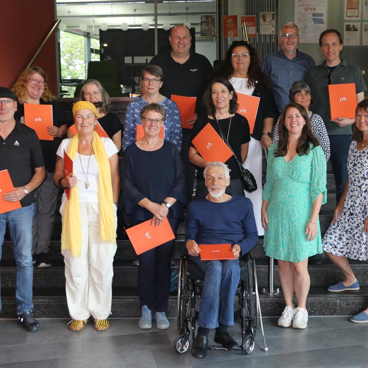 A group of 17 people is standing on a staircase, some holding orange documents. The participants are dressed in various styles and are smiling.