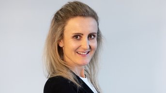 Eine Frau mit langen, blonden Haaren trägt einen schwarzen Blazer und steht mit verschränkten Armen. Sie lächelt leicht und blickt zur Seite. Der Hintergrund ist neutral und hell.