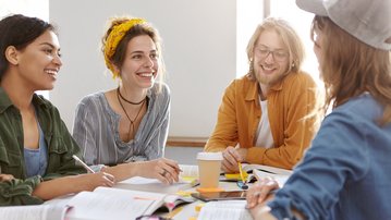 Vier junge Erwachsene sitzen an einem Tisch, lächeln und diskutieren, umgeben von Büchern und Notizen. Ein Kaffeebecher steht auf dem Tisch.