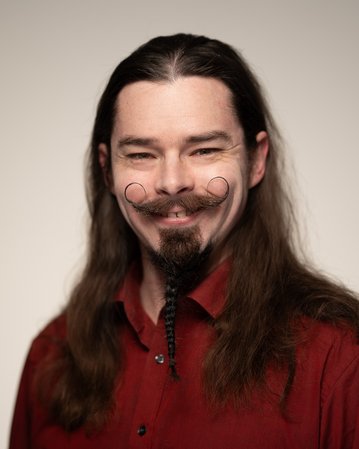 Ein Mann mit langen, dunklen Haaren und einem auffälligen Bart lächelt. Er trägt ein rotes Hemd und hat einen kunstvoll gestutzten Schnurrbart.