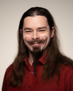 Ein Mann mit langen, dunklen Haaren und einem auffälligen Bart lächelt. Er trägt ein rotes Hemd und hat einen kunstvoll gestutzten Schnurrbart.