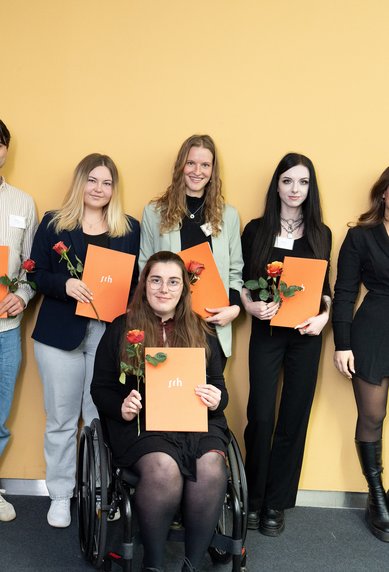 Gruppenzusammenstellung von sieben Personen, die Urkunden halten und Rosen tragen, vor einer gelben Wand. Ein Mann steht links.