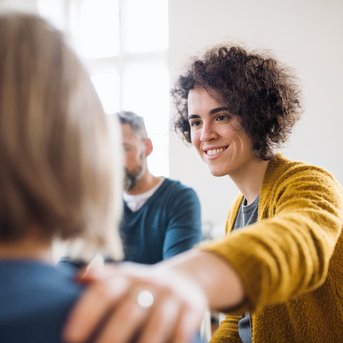 Eine Frau mit lockigem Haar lächelt und legt ihre Hand auf die Schulter einer anderen Person. Im Hintergrund sind zwei weitere Personen zu sehen, die an einem Gespräch teilnehmen.