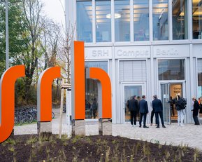A modern building with the inscription "SRH Campus Berlin" and a striking orange sculpture. People are standing in front of the entrance, chatting. Trees and bicycles are visible in the background.
