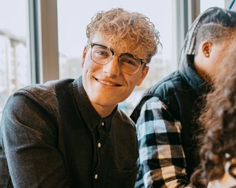 Junger Mann mit lockigem Haar und Brille, lächelt in einem Café, während er an einem Tisch sitzt. Im Hintergrund sind weitere Personen sichtbar.