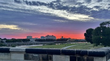Sunset over an urban landscape with clouds and a river in the foreground, surrounded by trees and meadows.