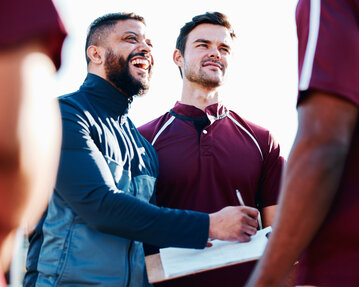 Two men in sportswear are standing together; one is holding a notebook and smiling while the other listens attentively.
