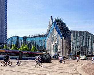 Moderne Architektur eines Gebäudes mit Glasfassade, umgeben von Radfahrern und Fußgängern. Im Vordergrund sind mehrere Fahrräder und Busse zu sehen.