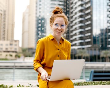 A woman with red hair and glasses is sitting outside in a yellow dress, working on a laptop in front of modern buildings.