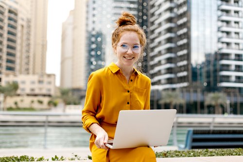 Eine Frau mit roten Haaren und Brille sitzt im Freien in einem gelben Kleid und arbeitet an einem Laptop vor modernen Gebäuden.