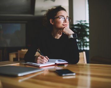 Eine nachdenkliche Frau mit Brille sitzt an einem Tisch, hält einen Stift und schreibt in ein Notizbuch. Auf dem Tisch liegen ein Laptop und ein Smartphone.