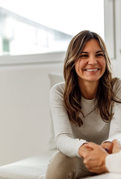 A smiling woman is sitting on a sofa, chatting with another person who is not visible in the picture.