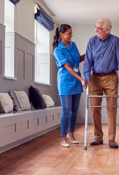 Eine Pflegekraft unterstützt einen älteren Mann mit einem Gehwagen in einem hellen Flur mit Sitzgelegenheiten.