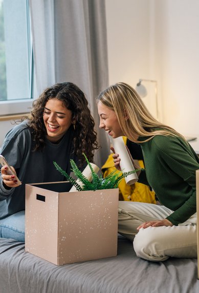 Two women are sitting on a bed in a bright room. One woman is holding a smartphone, while the other has a cup in her hand. Next to them are two moving boxes, one of which contains plants.