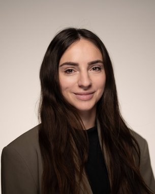 Eine Frau mit langen, braunen Haaren trägt einen Blazer und lächelt in die Kamera vor einem neutralen Hintergrund.