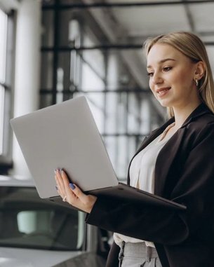 Eine junge Frau in einem Anzug steht in einem modernen Autohaus und arbeitet an einem Laptop, während sie lächelt. Im Hintergrund sind Autos und große Fenster zu sehen.