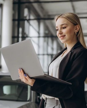 A young woman in a suit stands in a modern car dealership, working on a laptop while smiling. In the background, cars and large windows are visible.