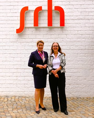 Zwei Frauen stehen vor einer weißen Wand mit dem roten Schriftzug "srh". Die Frau links trägt einen dunkelblauen Anzug mit einem bunten Schal, während die Frau rechts eine schwarz-weiße Jacke und schwarze Hose trägt.